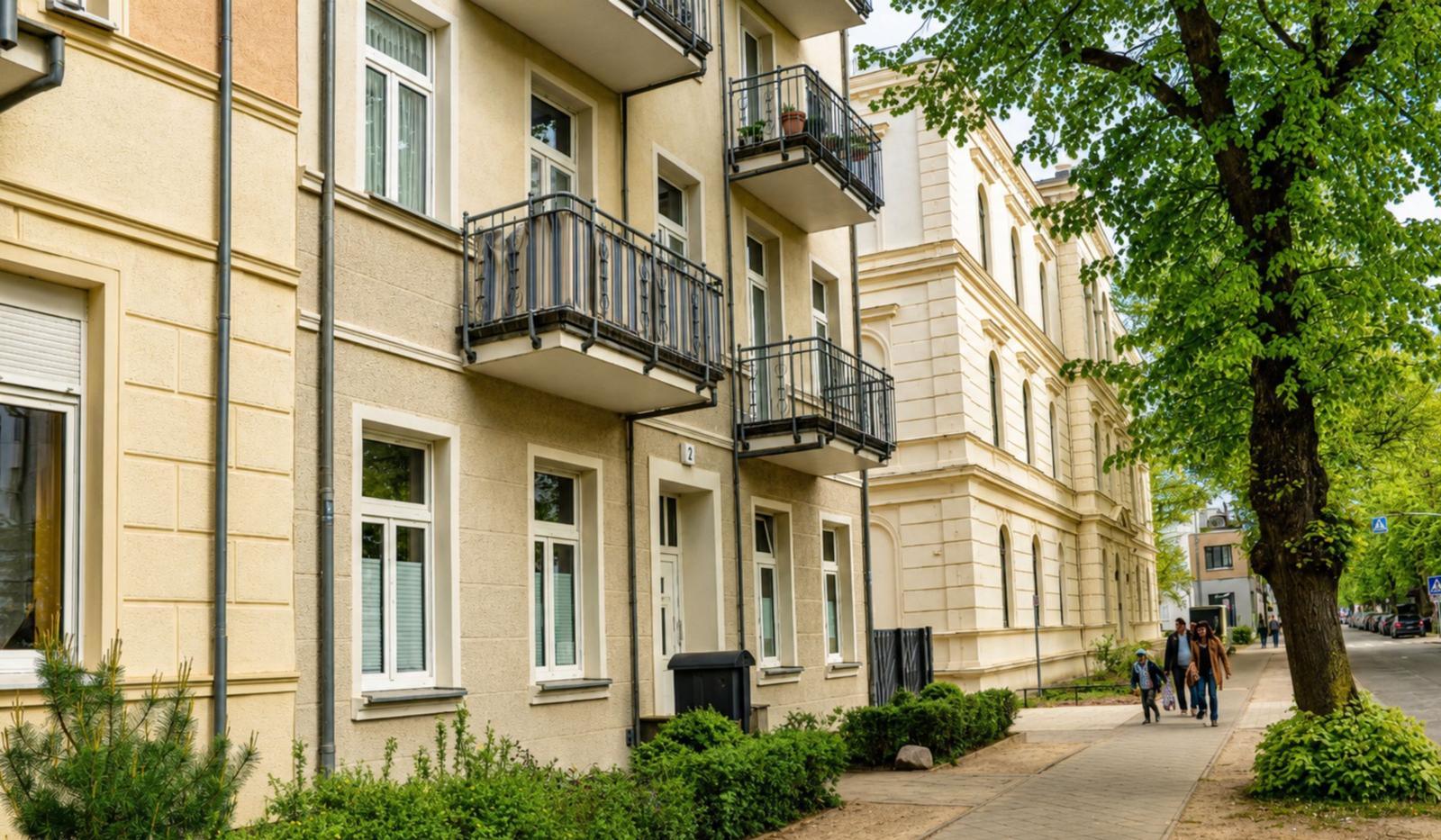 Haus mit Balkonen und grüner Straße. Menschen gehen auf dem Gehweg.