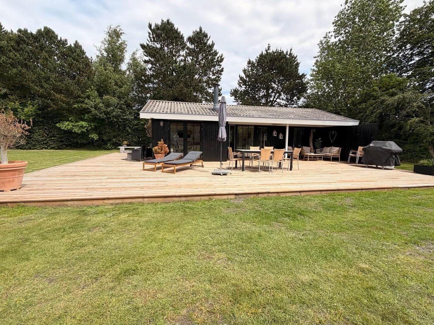 Haus mit großem Holzterrasse, Gartenmöbeln und Grillplatz. Umgeben von Bäumen und Grünfläche.