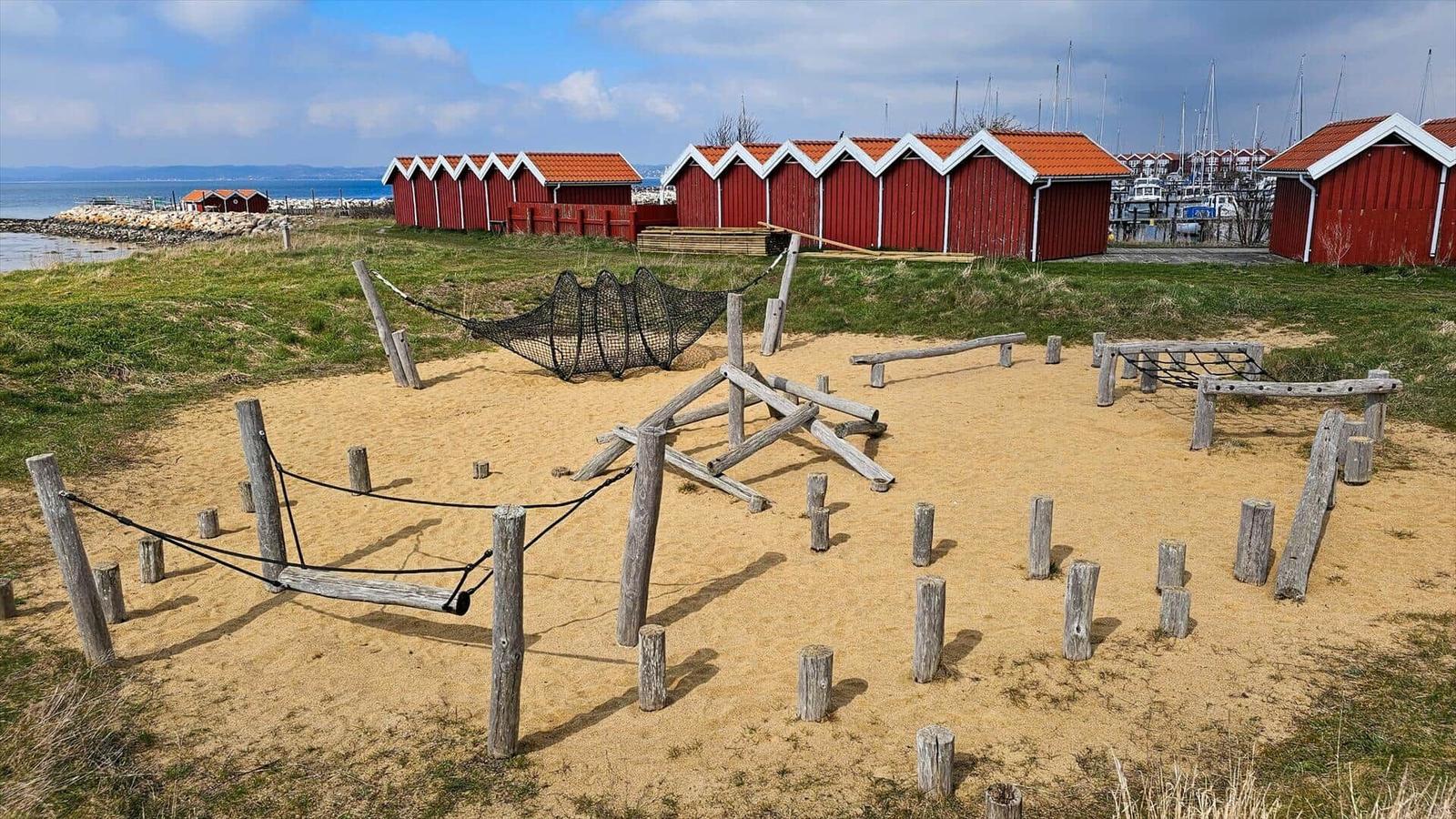 Sandplatz mit Holzspielgeräten und roten Strandhäusern im Hintergrund.
