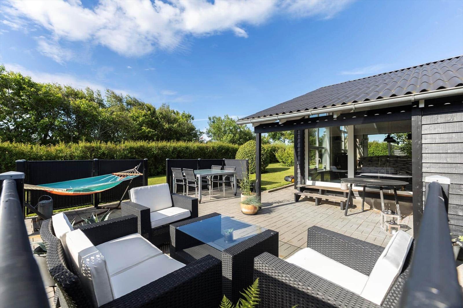 Terrasse mit Sitzgruppe, Tisch, Hängematte und Pflanzen. Hintergrund: Grünfläche und Bäume.