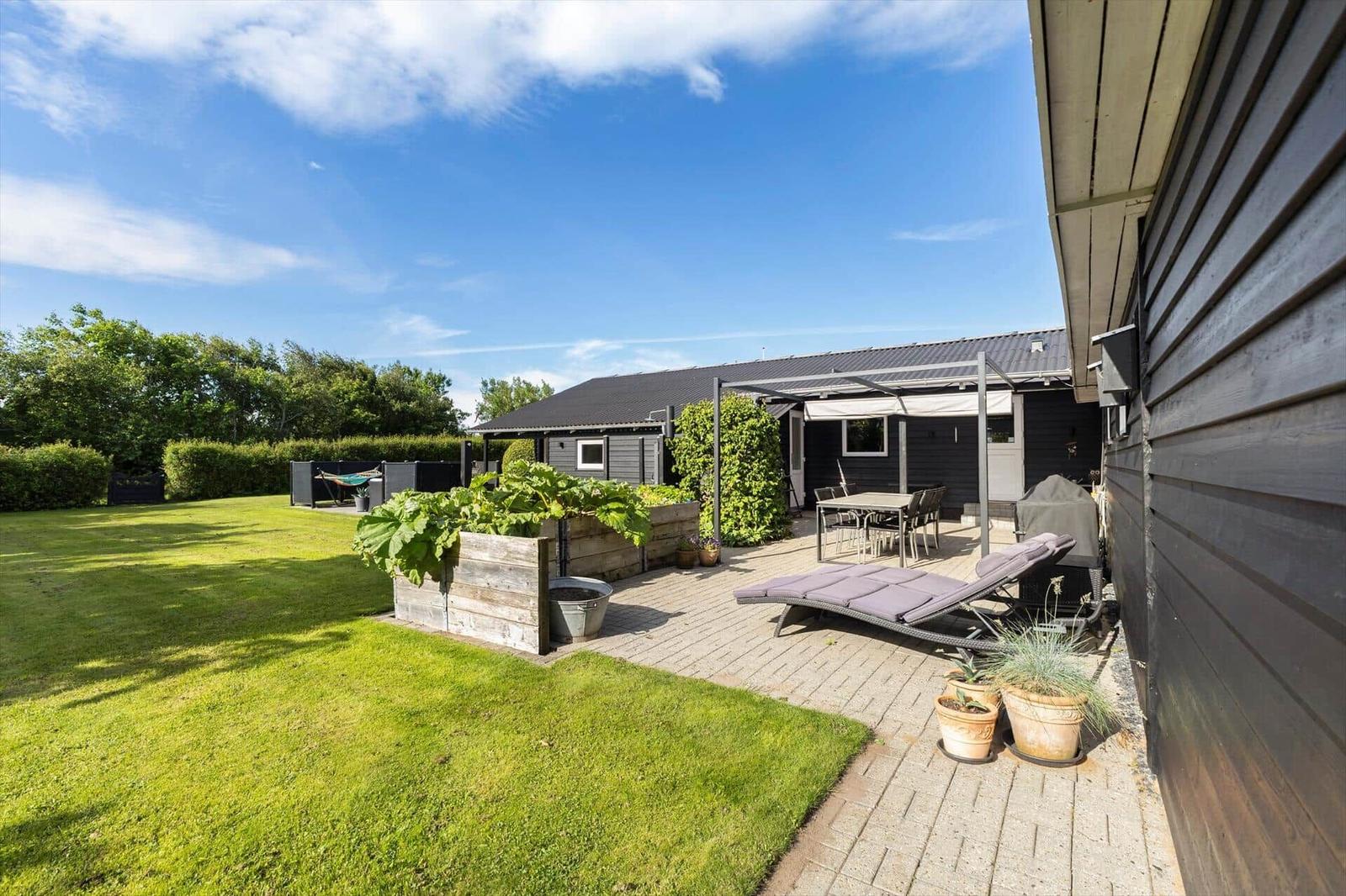 Terrasse mit Liegen, Tisch und Grill. Schwarze Fassade, Pflanzen und klarer Himmel.
