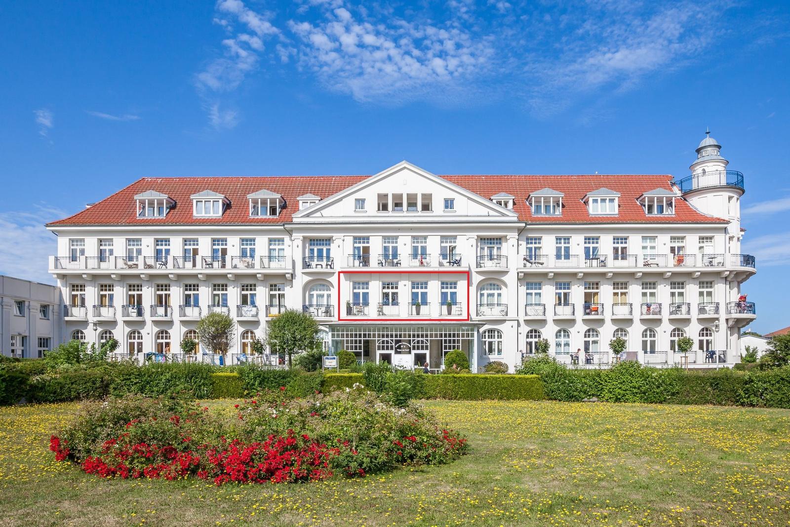 Großes weißes Gebäude mit rotem Dach und Balkonen. Garten mit Blumen vorne.