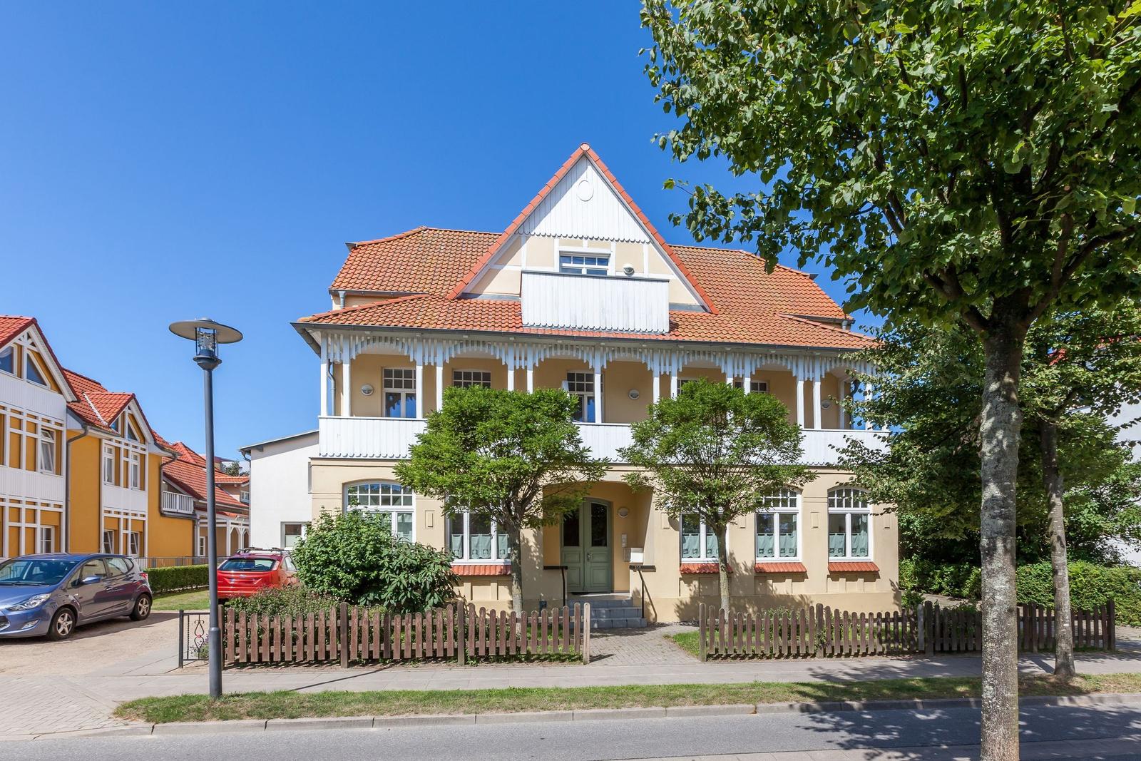 Haus mit rotem Dach und weißen Verandaelementen. Vorgarten mit Zaun und Bäumen.