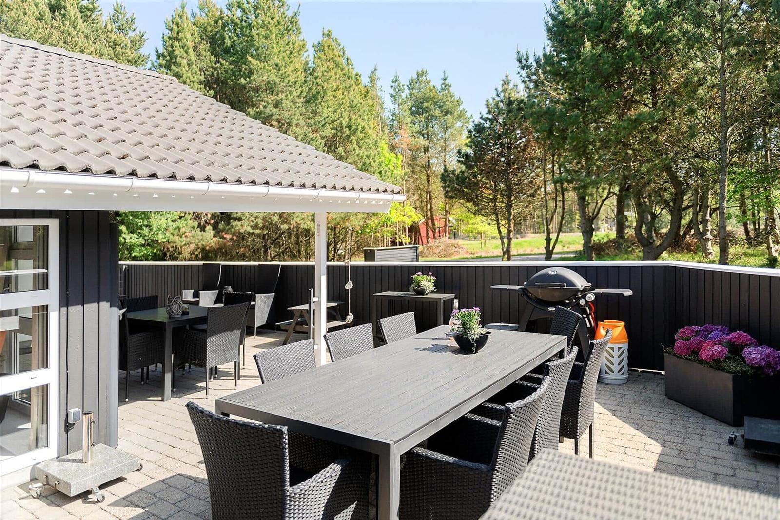 Terrasse mit Tisch, Stühlen, Grill und Pflanzen. Hintergrund: Wald und Zaun.