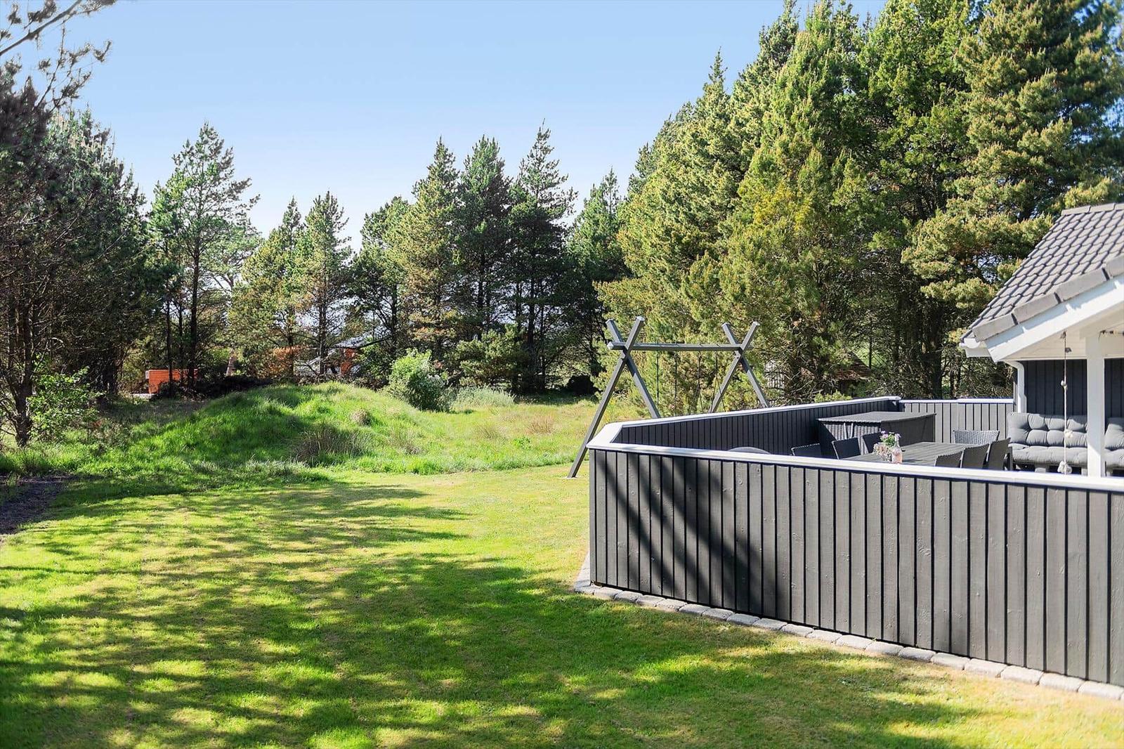 Garten mit grünem Rasen und Holzterrasse mit Sitzgelegenheiten. Hintergrund: Wald mit Nadelbäumen.