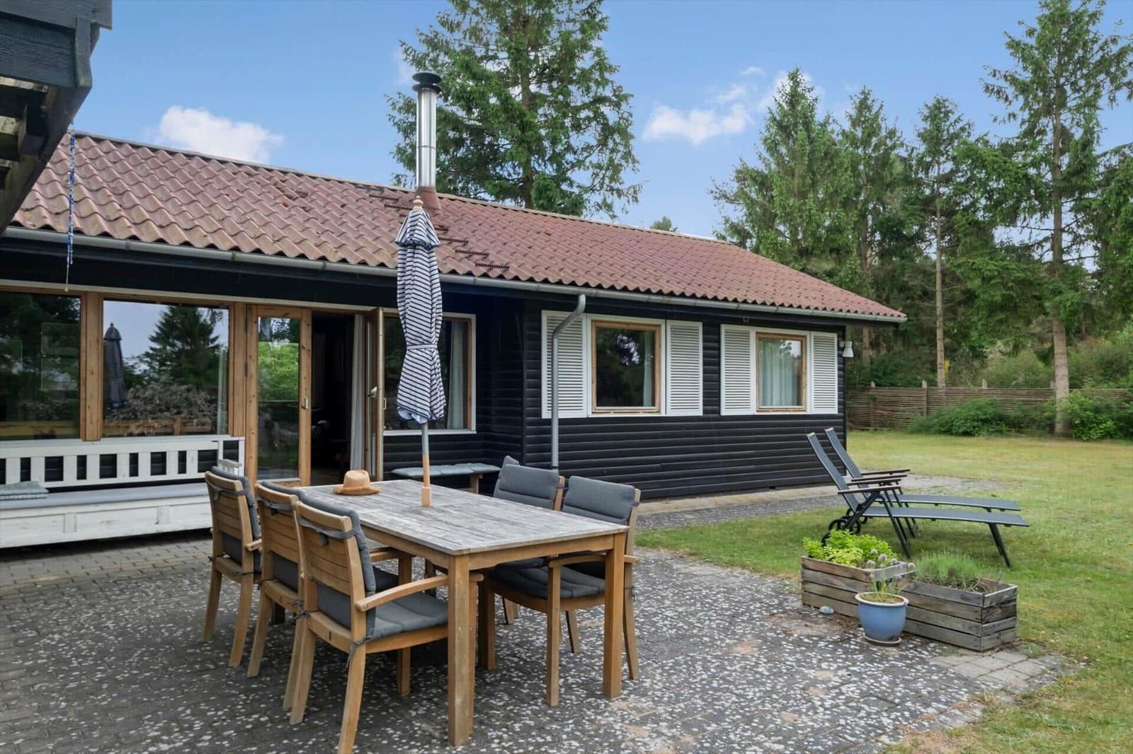 Terrasse mit Tisch, Stühlen und geschlossenem Sonnenschirm. Hintergrund: Grünfläche und Bäume.