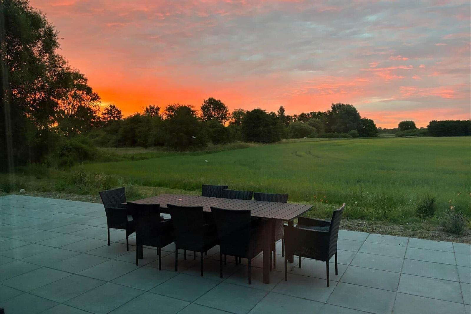Terrasse mit Tisch und Stühlen vor grünem Feld bei Sonnenuntergang.
