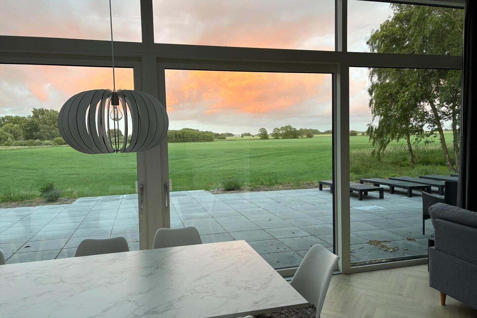 Großes Fenster mit Blick auf grünes Feld und Sonnenuntergang. Innen: Tisch, Stühle und Hängelampe.