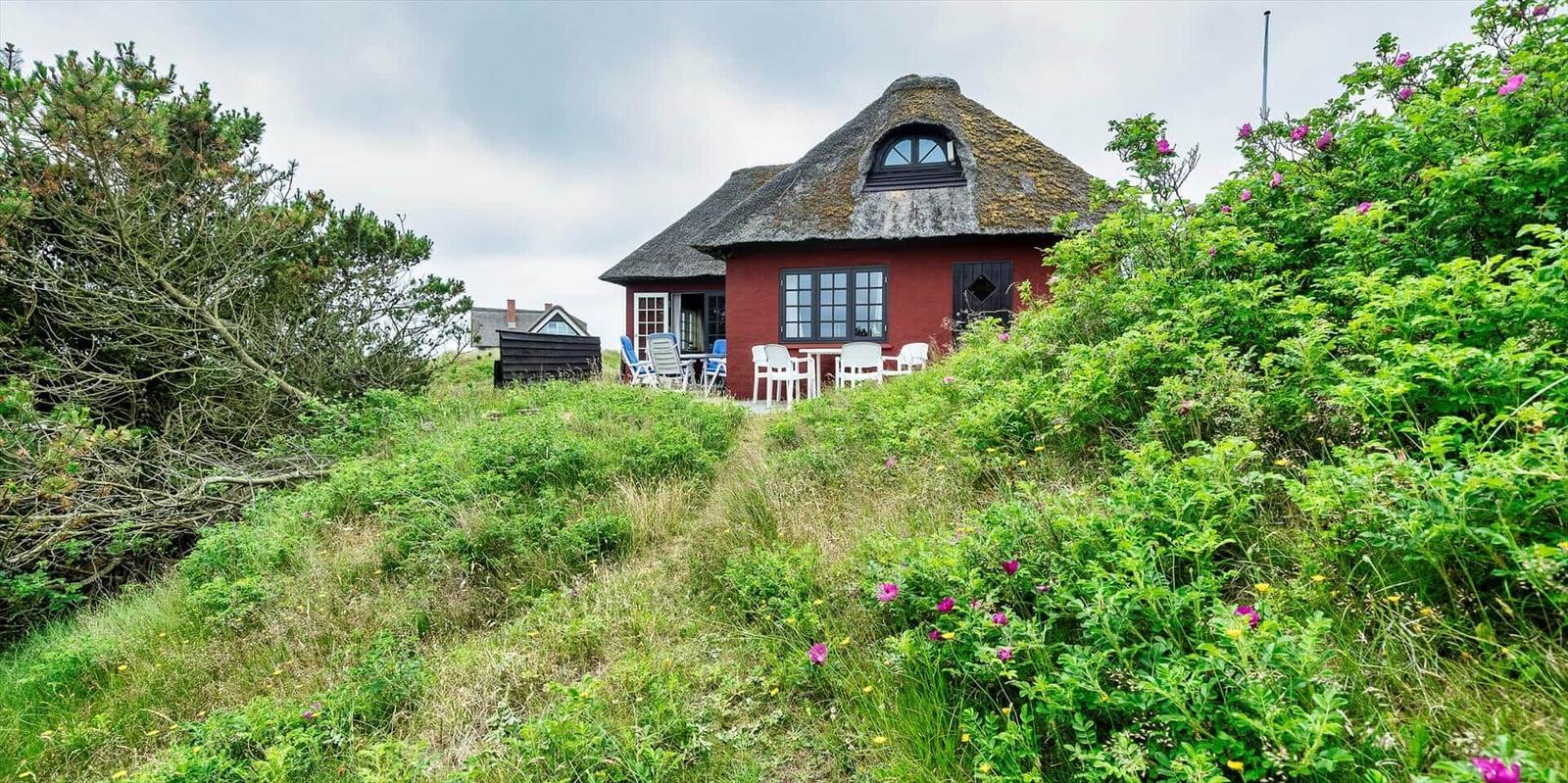 Rotes Haus mit Dach aus Stroh, Terrasse mit Tischen und Stühlen, umgeben von Grünflächen.