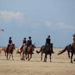 Fünf Reiter auf Pferden im Sand. Ein Kite fliegt im Hintergrund.
