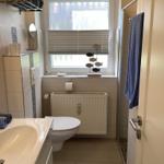 Badezimmer mit Toilette, Waschbecken und Dusche. Fenster mit Jalousie und Badewannenrand.