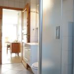 Badezimmer mit Dusche, Toilette und Waschbecken. Durchgang zu einem Raum mit Holztür.