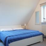 Schlafzimmer mit Dachschrägen, Bett mit blauer Bettwäsche, zwei Fenster und Spiegel an der Wand.