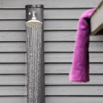 Außen-Dusche mit laufendem Wasser an grauer Wand, lila Handtuch hängt daneben.