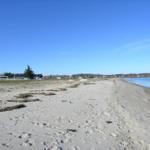 Strand mit Sand und Wasser, im Hintergrund Häuser und Bäume unter blauem Himmel.