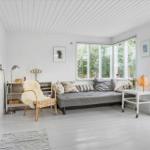 Wohnzimmer mit grauem Sofa, Wickerstuhl und großen Fenstern. Weiße Wände und Holzboden.