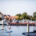 Zwei Personen stehen auf Stand-Up-Paddleboards auf dem Wasser vor einem Hafen.