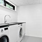 Waschraum mit Waschmaschine, Spüle und Kühlschrank. Weiße Wände und schwarzer Arbeitsplatz.