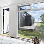 Schlafzimmer mit Blick auf Holzterrasse und grünen Garten. Weiße Wände und große Fenster.