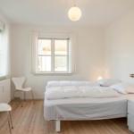 Schlafzimmer mit weißem Bett, zwei Fenstern und Holzboden.