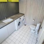 Waschraum mit Waschmaschine, Spülung und Toilette. Holzwand und Fliesenboden.