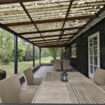 Terrasse mit Tisch und Stühlen unter einem Dach. Schwarze Holzwand und grüner Garten im Hintergrund.