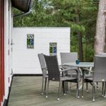 Terrasse mit Tisch und Stühlen, weißer Blockbau mit farbigen Fenstern, umgeben von Bäumen.