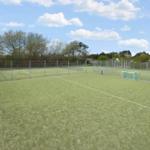 Green sports field with soccer goals and tennis net behind a fence.