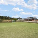 Green sports field with basketball hoops and goal. Buildings and trees in the background.