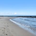 Strand mit Sand und sanften Wellen, Steine am Ufer, klarer Himmel mit wenigen Wolken.