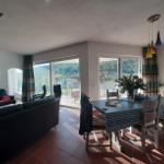 Wohn- und Essbereich mit Blick auf Garten und Hügel. Tisch, Stühle, Sofa und Fenster sind sichtbar.
