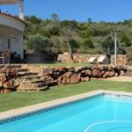 Pool mit Steinmauer, Terrasse und Garten im Hintergrund.