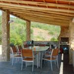 Außenbereich mit Tisch, Stühlen und Grillplatz. Steinpfeiler und Holzdecke.