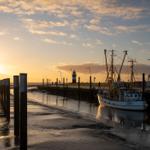 Fischerboot liegt im Hafen bei Sonnenuntergang. Leuchtturm im Hintergrund. Holzpfähle am Ufer.