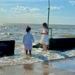 Zwei Kinder stehen am Ufer und spielen im Wasser. Im Hintergrund ist das Meer und ein Kitesurfer zu sehen.
