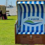 Strandkorb mit blau-weißem Muster und Logo auf grünem Rasen. Im Hintergrund See und Boot.