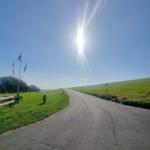 Sonniger Weg durch grünes Feld mit Parkplatz und Flaggenpfosten.