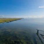 Wasserfläche mit Steinen und Holzbrücke im Vordergrund, Grasland und Windräder im Hintergrund.