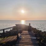 Eine Person sitzt auf einer Holzbrücke und blickt auf das Meer bei Sonnenuntergang.