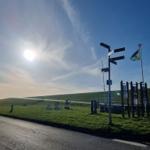 Sonniger Tag mit Wegweiser und Fahnenmast an einer grünen Hangstraße.