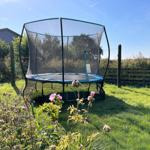 Trampoline with net in green garden with wooden fence and background vegetation.