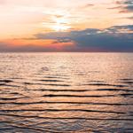 Sunset over calm water with gentle waves and clouds on the horizon.