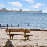 Holzbank am Strand mit Blick auf das Wasser und einen fernen Uferstreifen.
