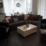 Wohnzimmer mit Sofa, zwei Sesseln und Couchtisch. Küchenbereich im Hintergrund.