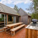 Holzterrasse mit Picknicktisch, Grill und Waschbecken. Glasfront zeigt Inneneinrichtung.