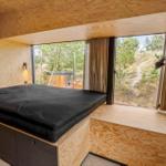 Schlafzimmer mit großem Fenster und Holzwandverkleidung. Blick auf Wald und Hügel.