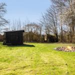 Grüner Garten mit Feuerstelle und kleinem Holzhaus. Bäume umgeben den Außenbereich.