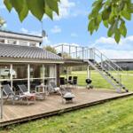 Haus mit Holzterrasse, Gartenmöbeln und Grillplatz unter blauem Himmel.