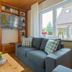 Ein Wohnzimmer mit grauem Sofa, Holztisch und TV. Fenster mit Blick auf Garten.