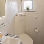 Badezimmer mit Spiegel, Waschbecken und Toilette. Fenster mit Pflanze und Handtuchhalter.